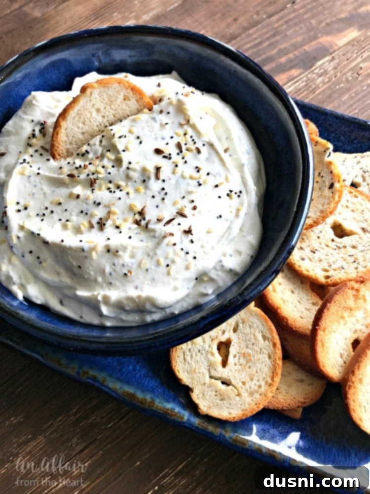 Whip Up Your Own Everything Bagel Dip 2 A vibrant, close-up shot of creamy Everything Bagel Dip in a rustic blue bowl, garnished generously with fresh chives and extra everything bagel seasoning. Bagel chips are perfectly arranged around the bowl, inviting you to dip.