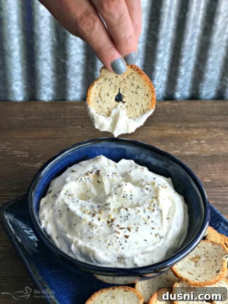 Whip Up Your Own Everything Bagel Dip 7 A close-up action shot of a crispy bagel chip being dipped into a bowl of creamy Everything Bagel Dip, showing the rich, thick texture of the dip clinging to the chip, ready for a bite.