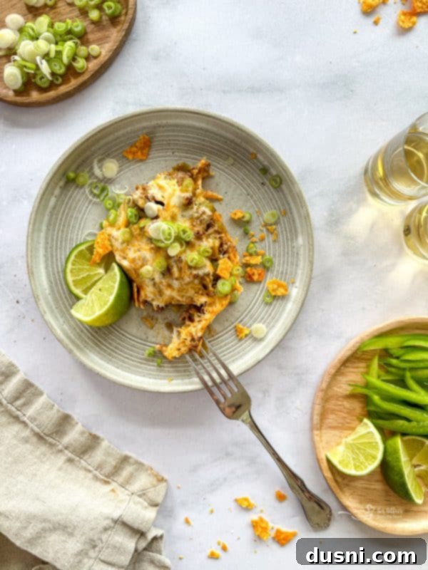 A serving of Mom's Doritos Taco Casserole in a square baking dish, with a portion scooped out, showcasing the distinct layers of crushed Doritos, seasoned ground beef, and melted cheese. A bowl of sour cream is visible in the background.