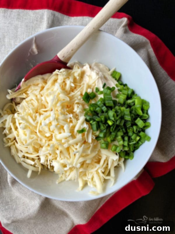 A hand mixing shredded Swiss cheese, mayonnaise, and sliced green onions in a large glass bowl with a spatula, illustrating the simple preparation process.