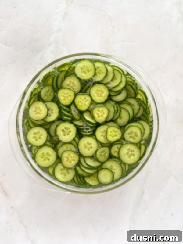 Close-up of a hand pouring ranch dressing over sliced cucumbers and onions in a bowl.
