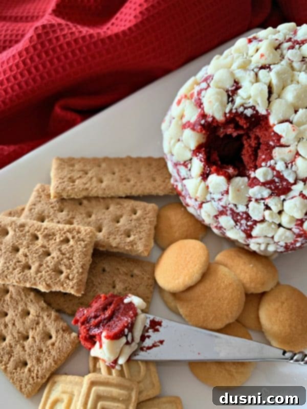Crimson Velvet Cheese Bomb 2 Red Velvet Cheese Ball, a delicious dessert with a red hue and white chocolate chip coating, on a serving platter with a spreader.