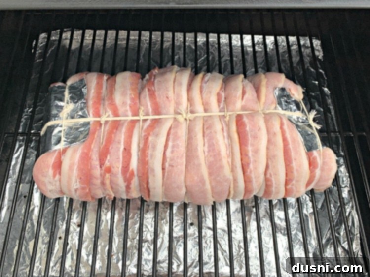 Pastırmalı Lahana Turşusu Dolgulu Domuz Kontrafile 12 Bacon-wrapped pork loin placed on a smoker grill with a fresh drip pan underneath.