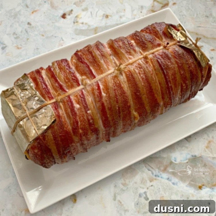 Pastırmalı Lahana Turşusu Dolgulu Domuz Kontrafile 14 Cooked bacon-wrapped pork loin on a white platter, glistening and ready to be carved.