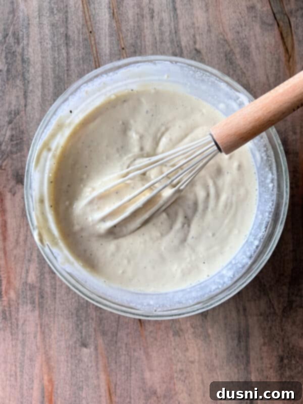 A whisk blending mayonnaise, dill pickle juice, and black pepper in a glass bowl to create the creamy dressing.