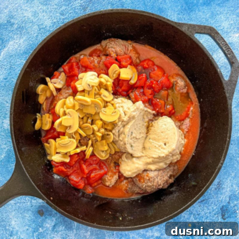 Overhead view of pepper steak simmering in a pan with mushrooms, tomatoes, and a creamy sauce, cooking gently to meld flavors.