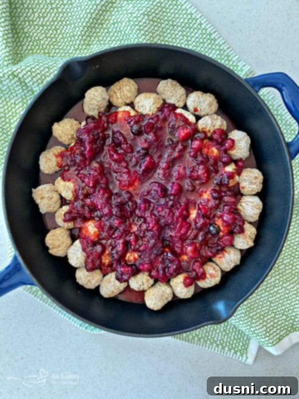 Dorothy Cranberry Meatballs 6 Hands mixing the sauce for Dorothy's Cranberry Meatballs in a clear glass bowl, showcasing the vibrant red color of the combined ingredients.