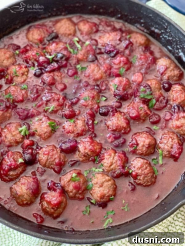 Dorothy Cranberry Meatballs 7 Dorothy's Cranberry Meatballs simmering in a cast iron skillet on the stovetop, coated in a thick, glistening red sauce, indicating they are almost ready.
