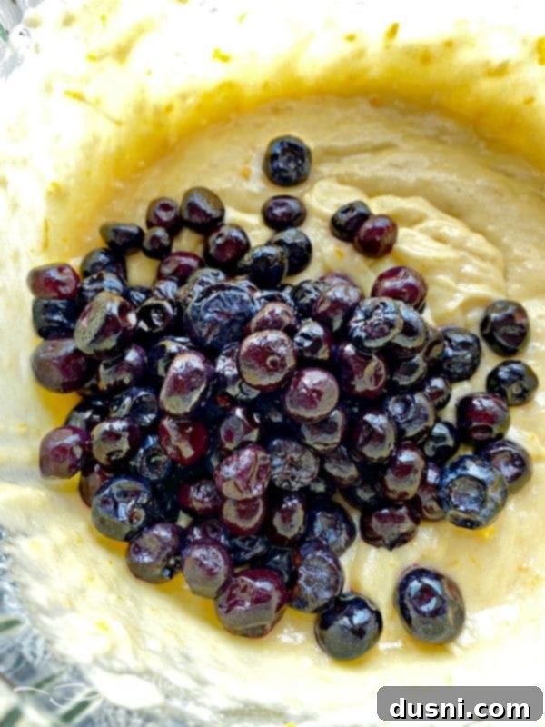 Blueberries being gently folded into a light-colored quick bread batter in a glass bowl.