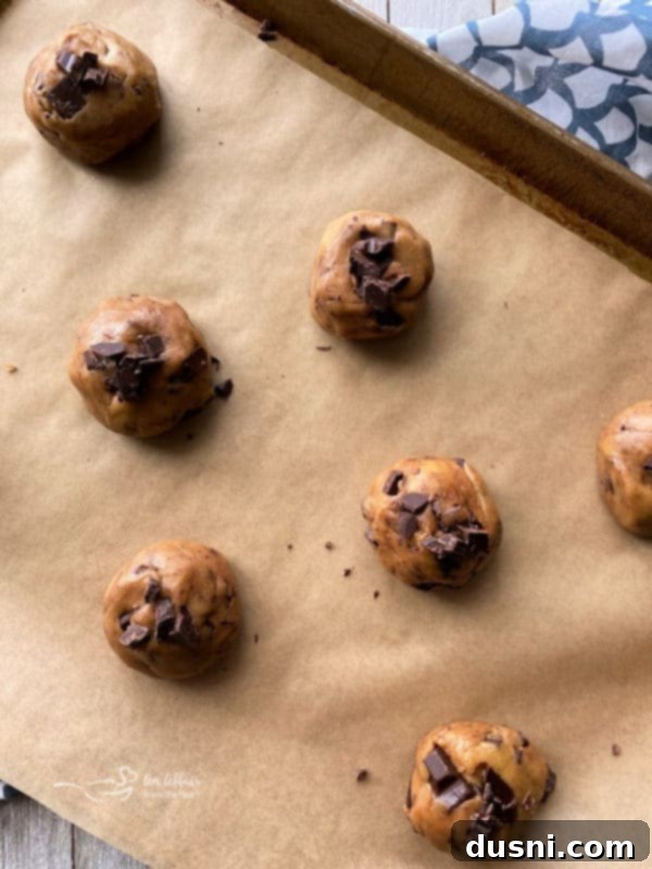Scoops of brown butter chocolate chunk cookie dough, topped with extra chocolate pieces, ready for baking on a parchment-lined tray.