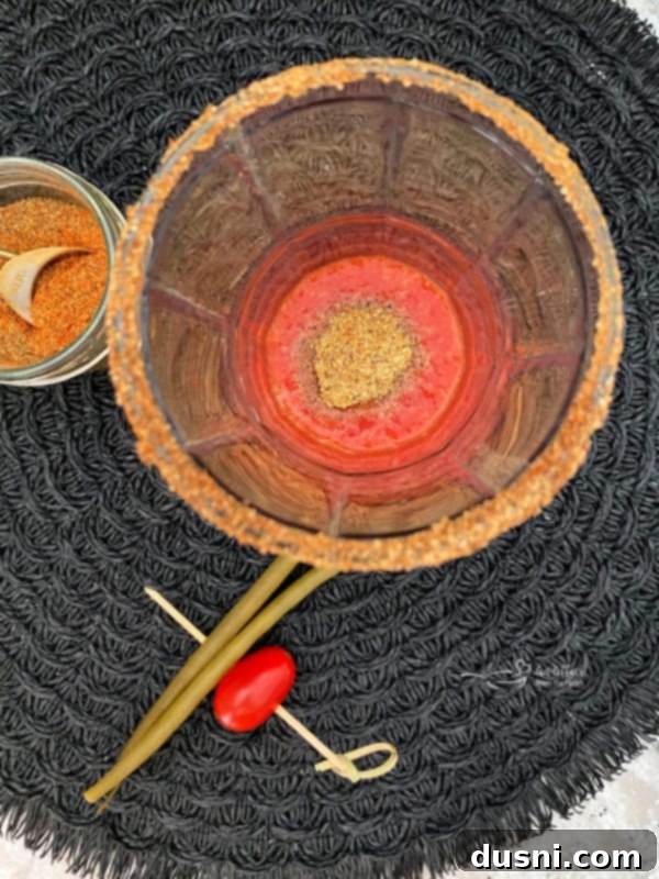A small spoonful of the homemade Bloody Mary Dry Spice blend being added to the tomato juice in a glass.