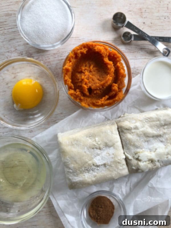 Ingredients laid out for pumpkin pie turnovers: pumpkin puree, puff pastry, egg, sugar, cream, pumpkin pie spice, powdered sugar, butter, milk