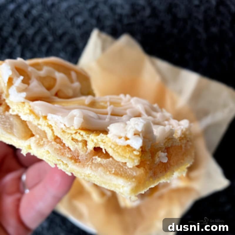 Effortless Apple Crumble Bars 6 Close-up of a square Apple Bar, showcasing the golden-brown flaky crust, tender cinnamon apples, and sweet white glaze, on a white plate with a fork.