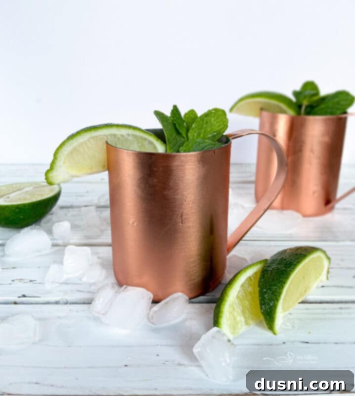 The Ginger Kick from Moscow 8 Two copper mugs with Moscow Mules, garnished with lime and mint, on a wooden surface.