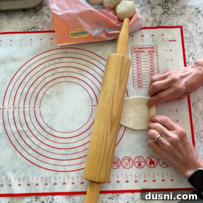 A frozen dinner roll being flattened into an oval shape with a rolling pin on a floured surface.