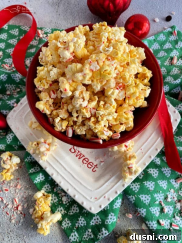 Peppermint Bark Popcorn Delight 2 A large bowl of white peppermint bark popcorn, with a few candy canes nestled in the popcorn. The popcorn is covered in white coating and crushed red peppermint pieces.