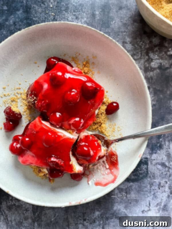 A slice of no-bake cherry dessert on a white plate, showcasing the graham cracker crust, creamy white filling, and bright red cherry topping.