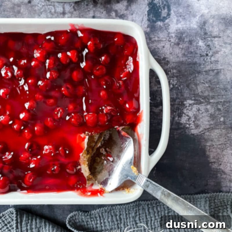 Close-up of the cherry dessert showing the layers: graham cracker crust, creamy white filling, and bright red cherry topping, ready for serving.