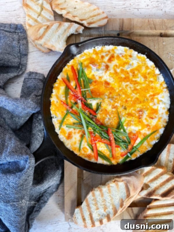 A cast iron skillet filled with Slow Cooker Crab Dip, garnished with green onions and red peppers, served with toasted baguette slices.