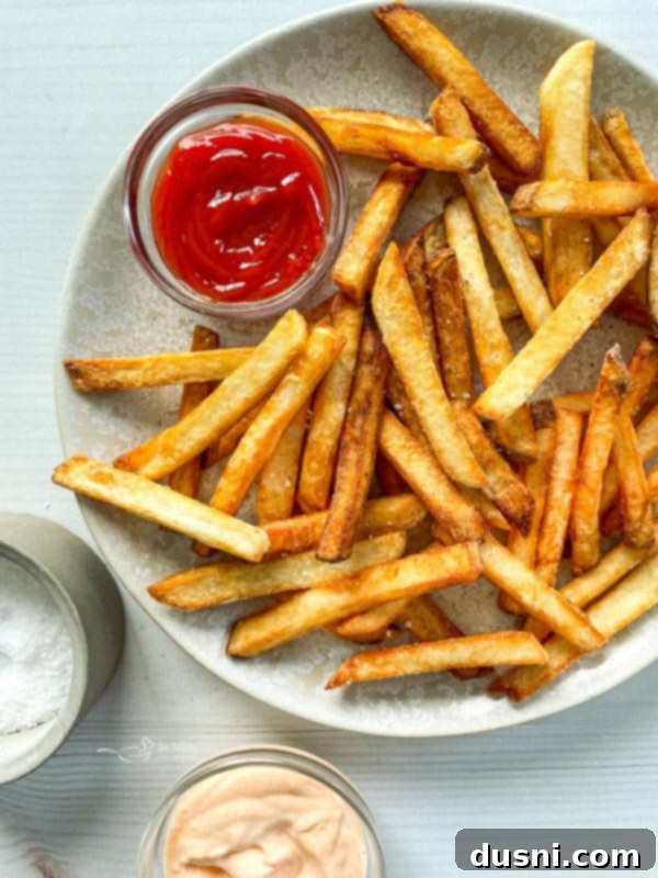 Top view of homemade double fried French fries on white plate with ketchup and salt, showcasing their golden crispiness.
