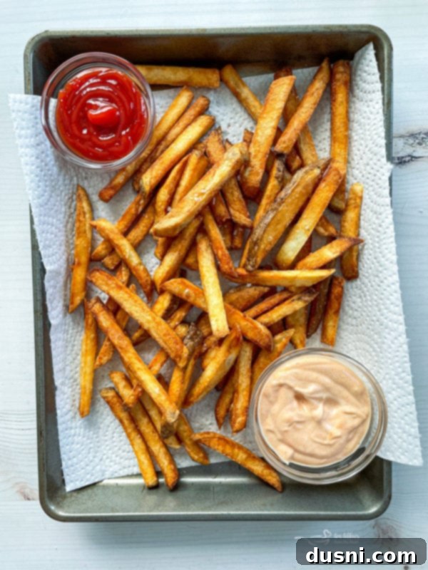 Homemade double fried french fries on baking sheet with parchment paper, with ketchup and sauce.