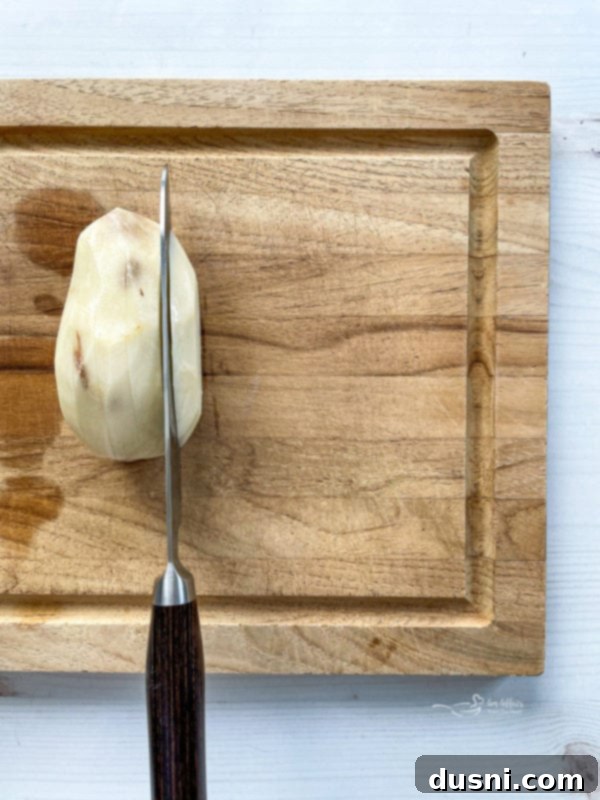 One peeled potato being sliced on wooden cutting board, demonstrating the initial preparation step.