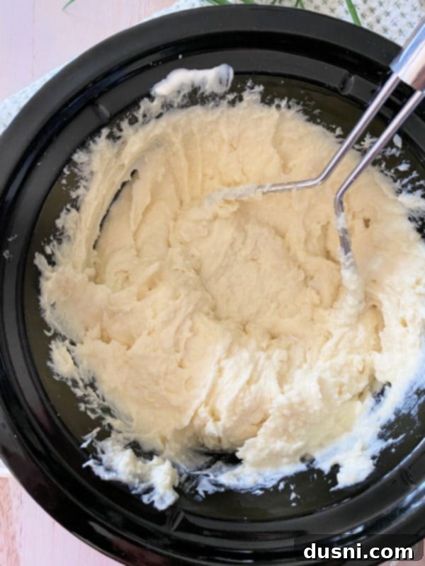 Ultimate Creamy Slow Cooker Mashed Potatoes 13 Creamy mashed potatoes in the Crockpot with a potato masher, showing smooth consistency