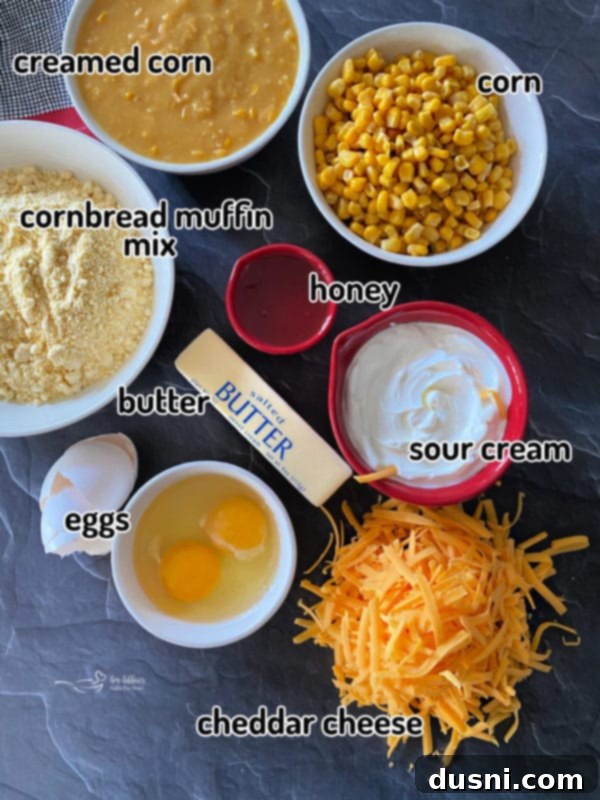 Individual ingredients for corn pie casserole laid out: creamed corn, corn muffin mix, butter, eggs, shredded cheese, sour cream, and honey on a gray surface