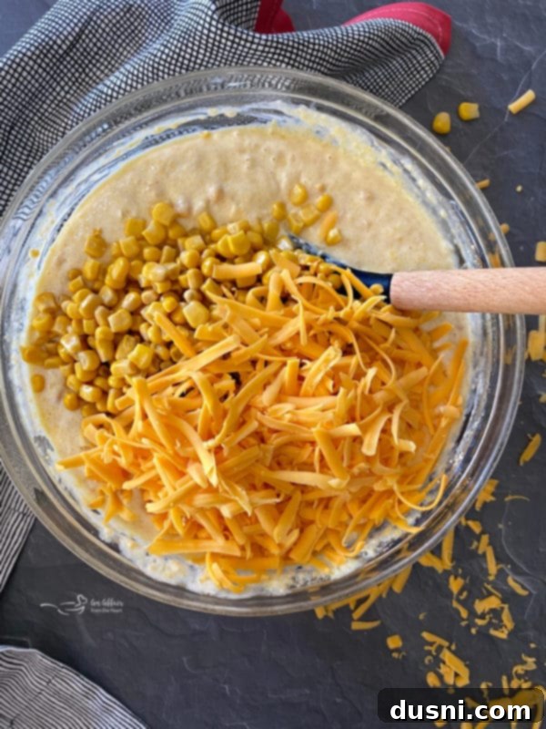 Corn pie casserole mixture in a bowl with shredded cheddar cheese and drained whole corn kernels, ready to be folded in