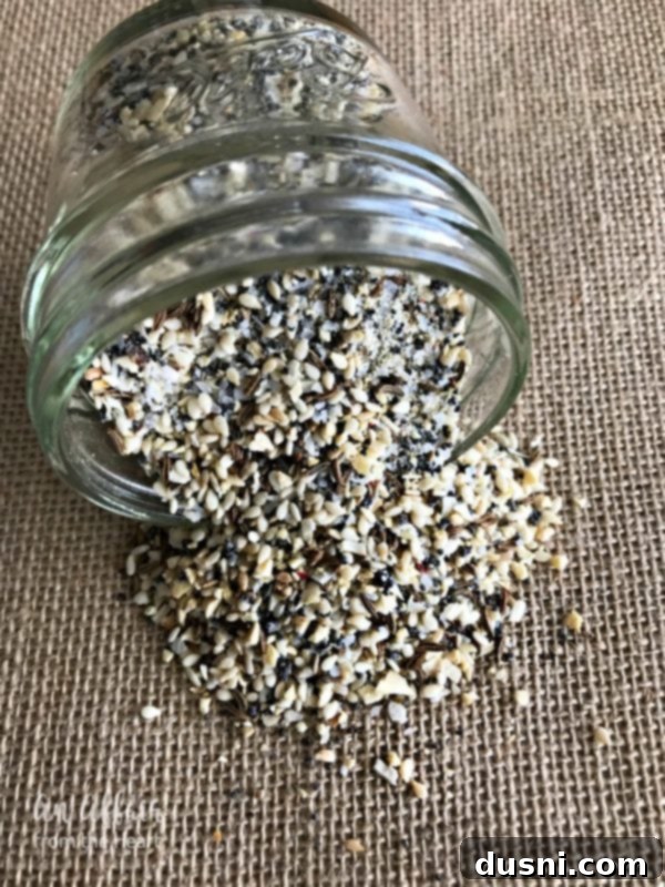 Everything Bagel Spice spilling out of a glass jar, showing sesame seeds, poppy seeds, garlic, onion, and salt.