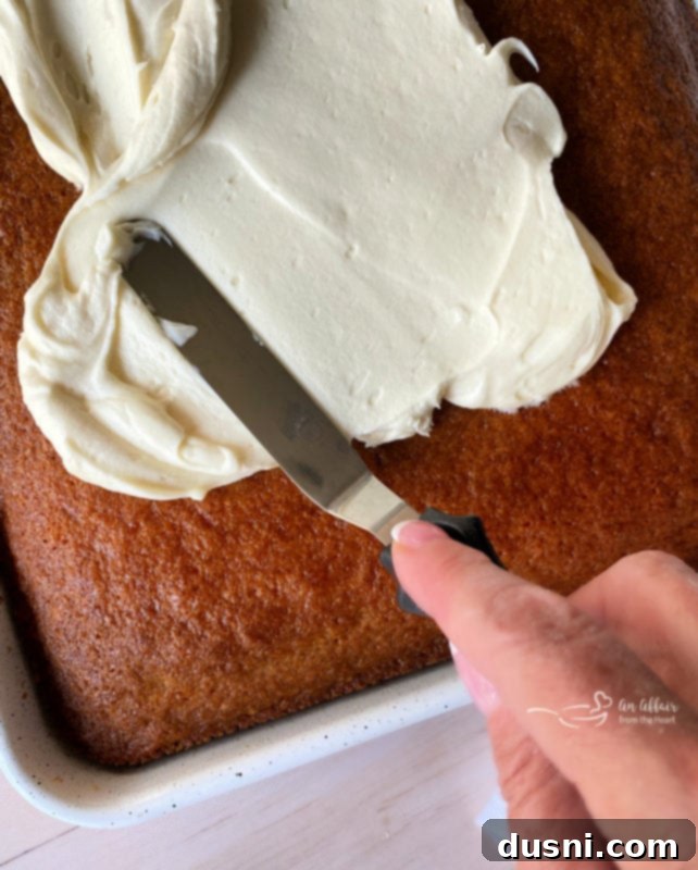 Carrot Cake Bars Recipe A hand gently spreading a generous layer of smooth cream cheese frosting over the cooled carrot cake using an offset spatula, ensuring even coverage.