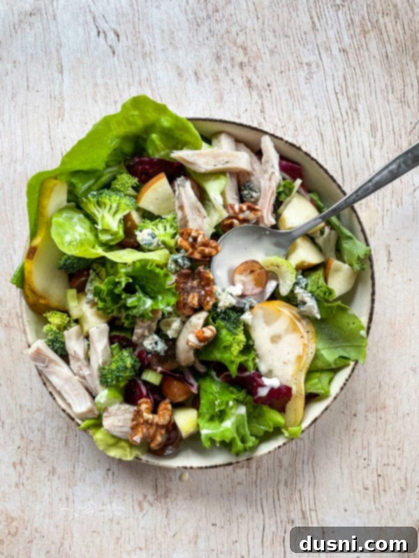 Waldorf Salad A close-up, top-down shot of a Grilled Chicken Waldorf Salad, beautifully presented in a white bowl with a spoon, highlighting the textures of chicken, walnuts, grapes, and greens.