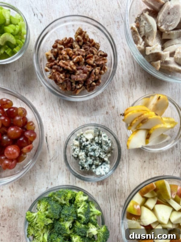 Waldorf Salad Recipe with Chicken A vibrant selection of fresh Waldorf salad ingredients including broccoli, blue cheese crumbles, green and red grapes, sliced pears, toasted walnuts, crisp celery, and grilled chicken slices, artfully arranged on a rustic wooden surface.