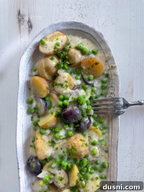 A white serving dish presenting cooked new potatoes and peas with creamy Bechamel sauce, garnished with fresh chives.
