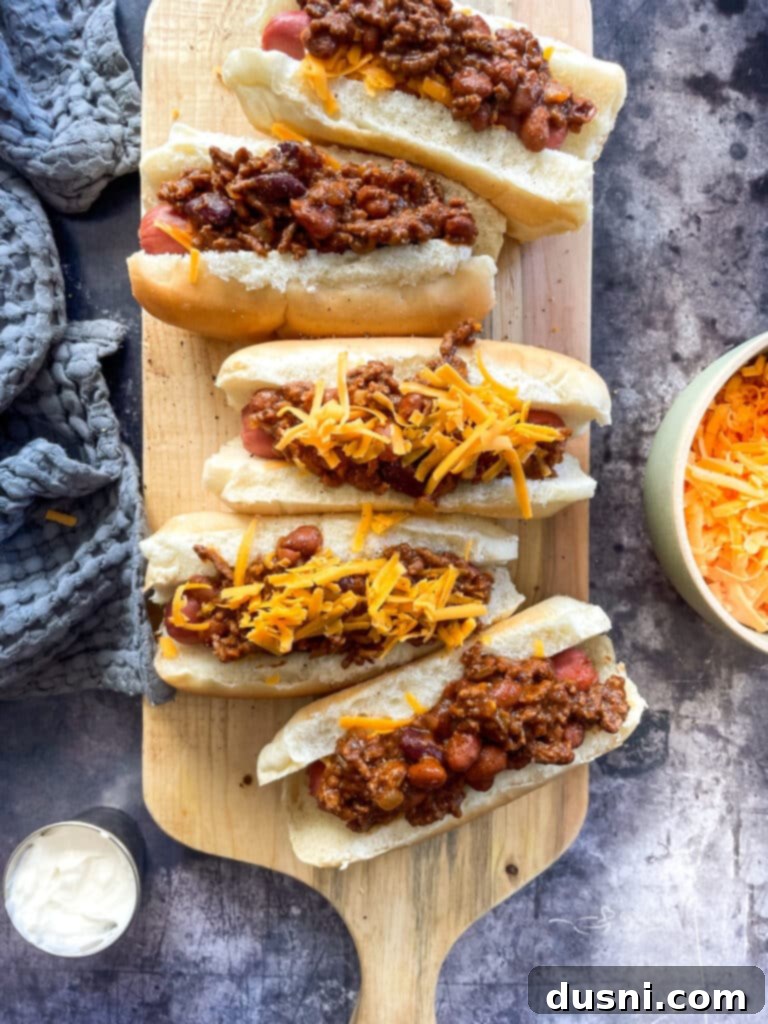 Hearty Slow Cooker Buffalo Chicken Chili 13 Hands placing shredded cheese on top of two chili dogs, with visible chili and hot dogs.