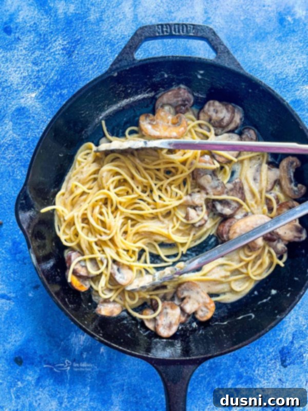 Cooked spaghetti noodles tossed with sautéed mushrooms in a skillet, creating a delicious creamy mushroom pasta.