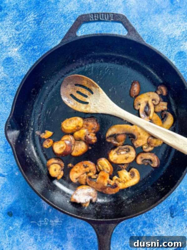 Sliced mushrooms cooking in a skillet with butter and olive oil, releasing their moisture and browning.