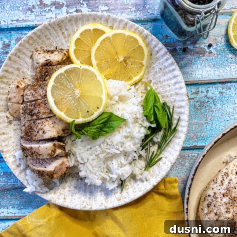 A pristine white plate holds a perfectly cooked lemon pepper chicken breast, garnished with fresh lemon wheels and basil, served alongside fluffy rice.