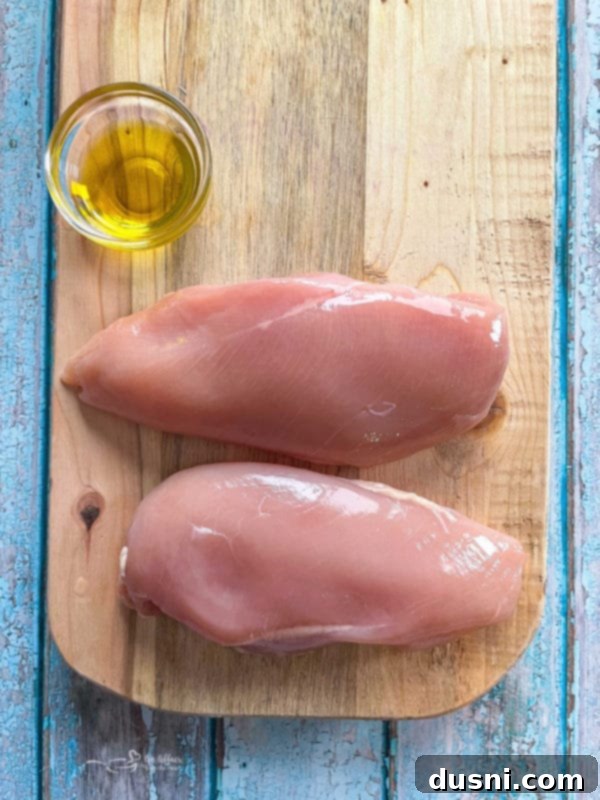 Two raw chicken breasts on a cutting board, brushed with olive oil, ready for seasoning and baking.
