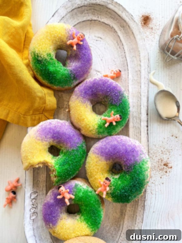 Close-up of fluffy baked king cake donuts decorated with purple, green, and gold sugar, with a small plastic baby on top of one donut.