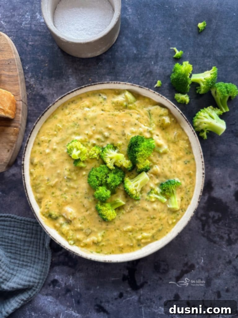 Two bowls of creamy broccoli cheese soup on a wooden table, with fresh broccoli florets and a spoon.