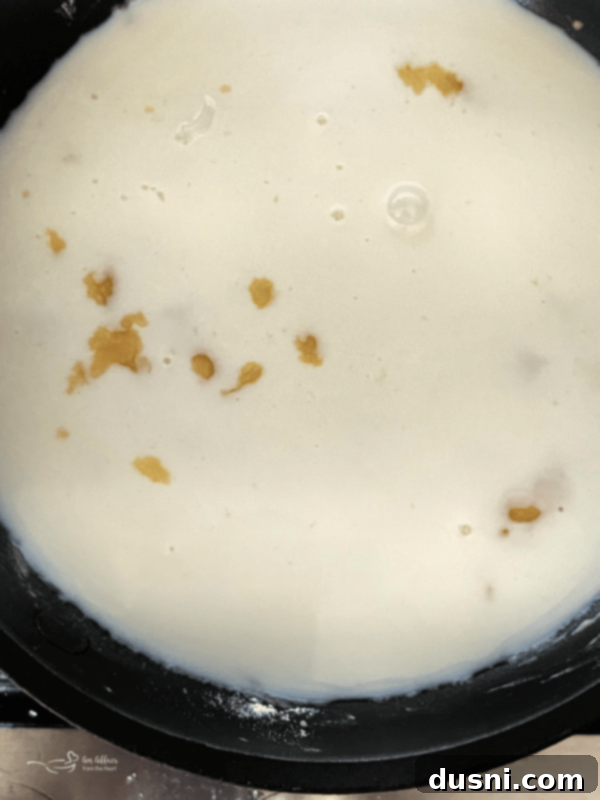 Milk being slowly poured into the roux in a skillet while being whisked.