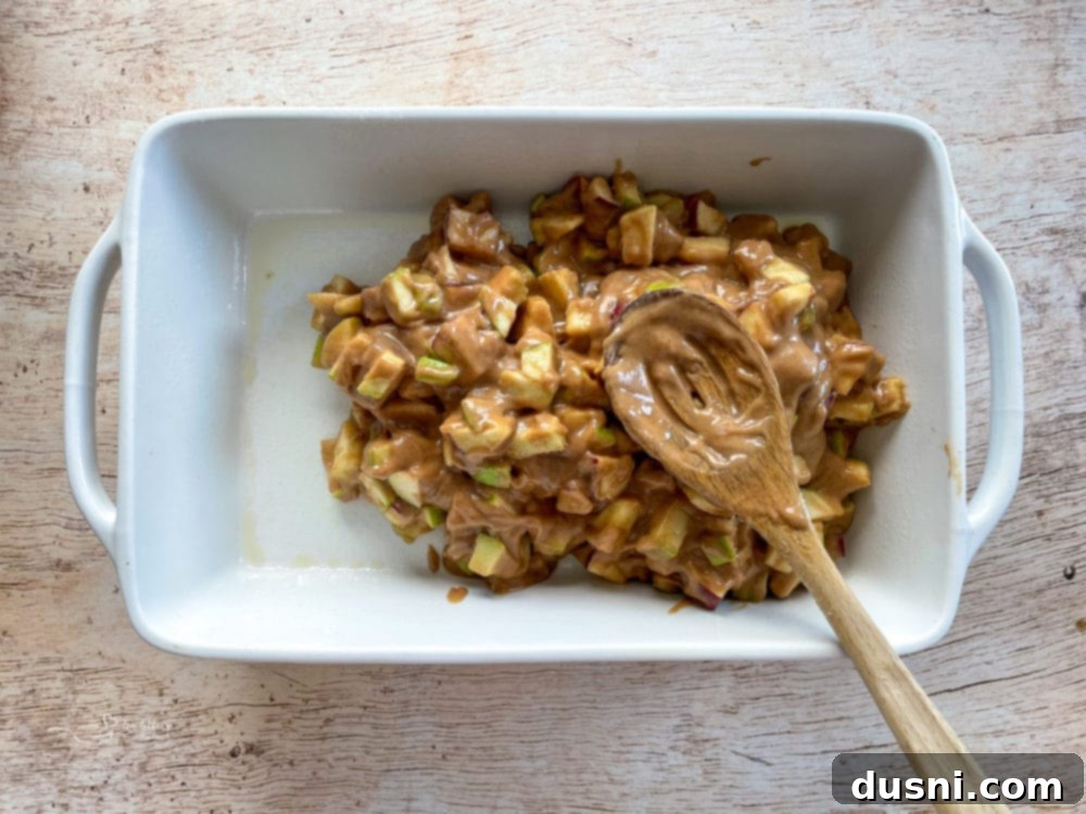 The finished apple cake batter being spooned into the prepared baking dish with a wooden spoon, ready for the oven.