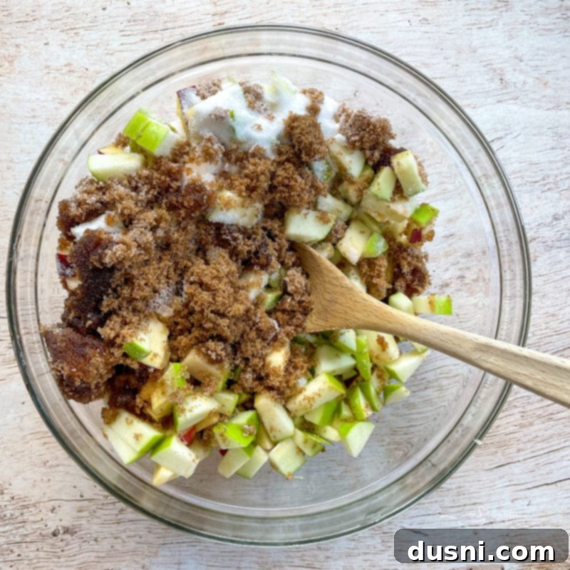 Diced apples mixed with both granulated and brown sugar in a large bowl, glistening and ready for the next ingredients.