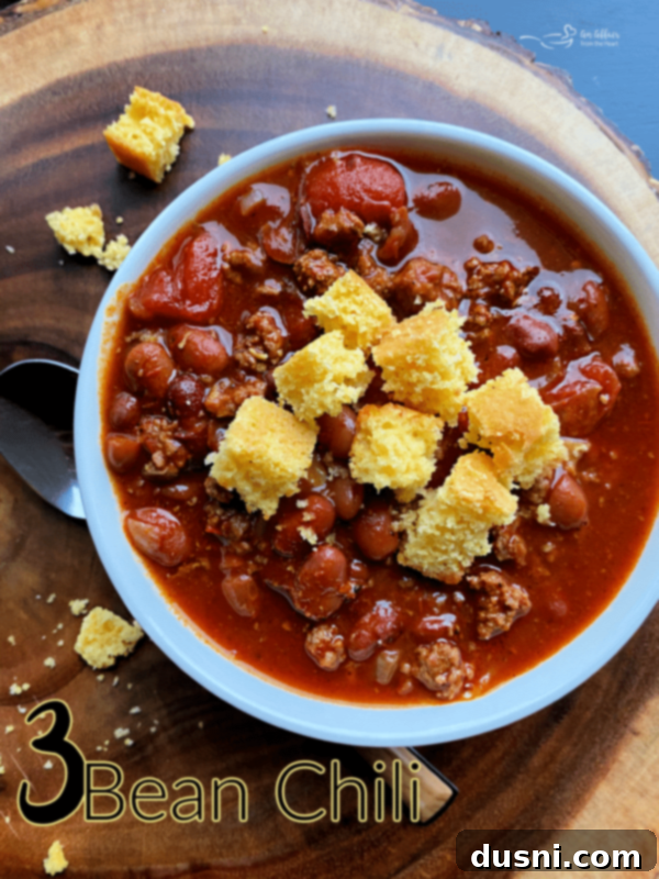 A bowl of Three Bean Chili garnished with cornbread, sour cream, and fresh herbs, presented on a light background, emphasizing its homemade appeal.