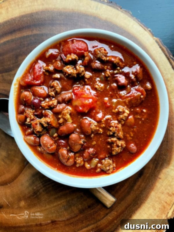A large bowl of Three Bean Chili, topped with shredded cheddar cheese, a dollop of sour cream, and a sprig of cilantro, against a dark, cozy background.