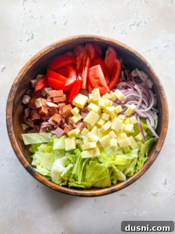 Adding chopped vegetables and meats to the lettuce base in a large mixing bowl.