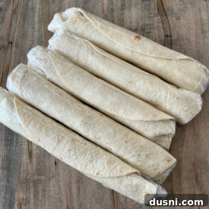Image showing a Reuben Style Tortilla Roll-Up tightly wrapped in plastic wrap, ready for chilling.