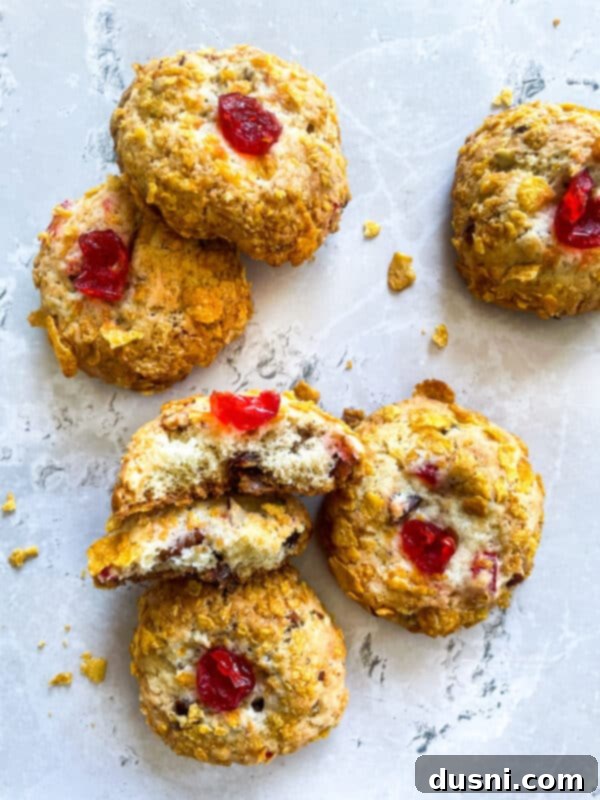 A batch of unbaked Cherry Wink Cookies, rolled in cornflakes and topped with cherries, on a baking sheet.