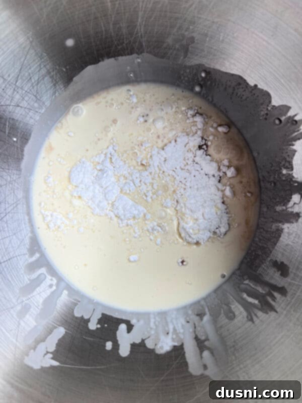 Close-up of heavy whipping cream, powdered sugar, and vanilla being whipped in a stand mixer, creating soft peaks.